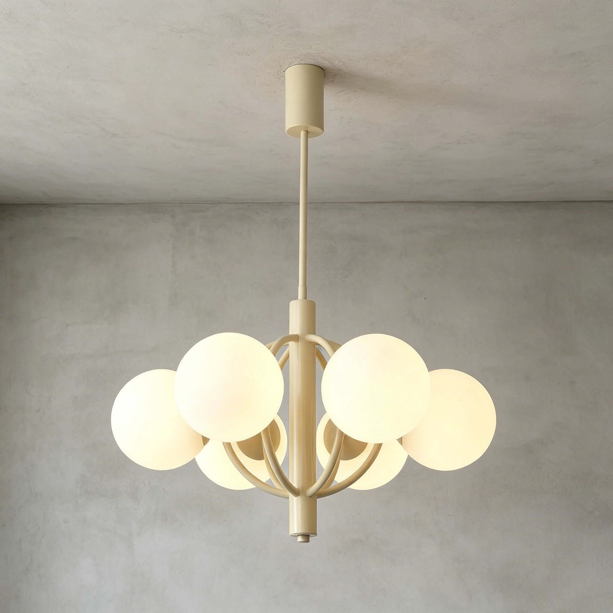 A Clérachic chandelier with milk-white globes and curved beige arms, lit and hung against a grey concrete wall.
