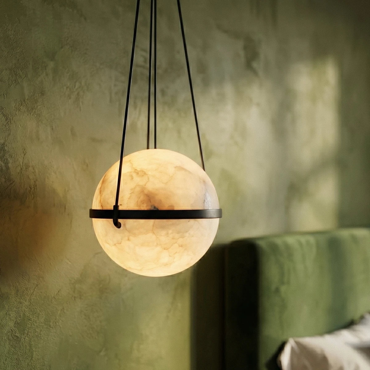 Clérachic close view of a lit alabaster globe with a dark metal band and cords, against green limewash and a velvet headboard.