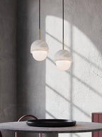 A pair of Clérachic Mini Alabaster Globe Pendants hung over a stone café table against a sunlit concrete wall.