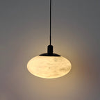The Clérachic Mini Discus pendant light switched on, alabaster glowing honey-cream and washing warm light across a plain wall.
