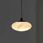Clérachic Mini Discus pendant light lit against a dark grey wall, cloudy veining surfacing through the warm stone shade.