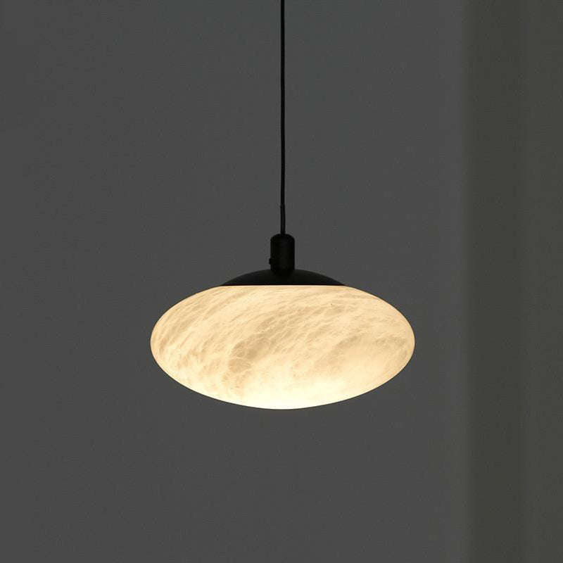 Clérachic Mini Discus pendant light lit against a dark grey wall, cloudy veining surfacing through the warm stone shade.