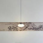Clérachic Mini Discus pendant light hanging low over an ink-wash landscape panel, one small glowing accent in a wide room.