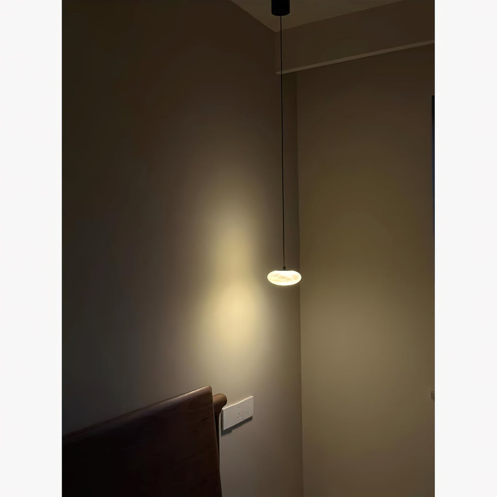 Clérachic Mini Discus pendant light on a long drop beside a bed, pooling soft amber light down a dim bedroom corner.