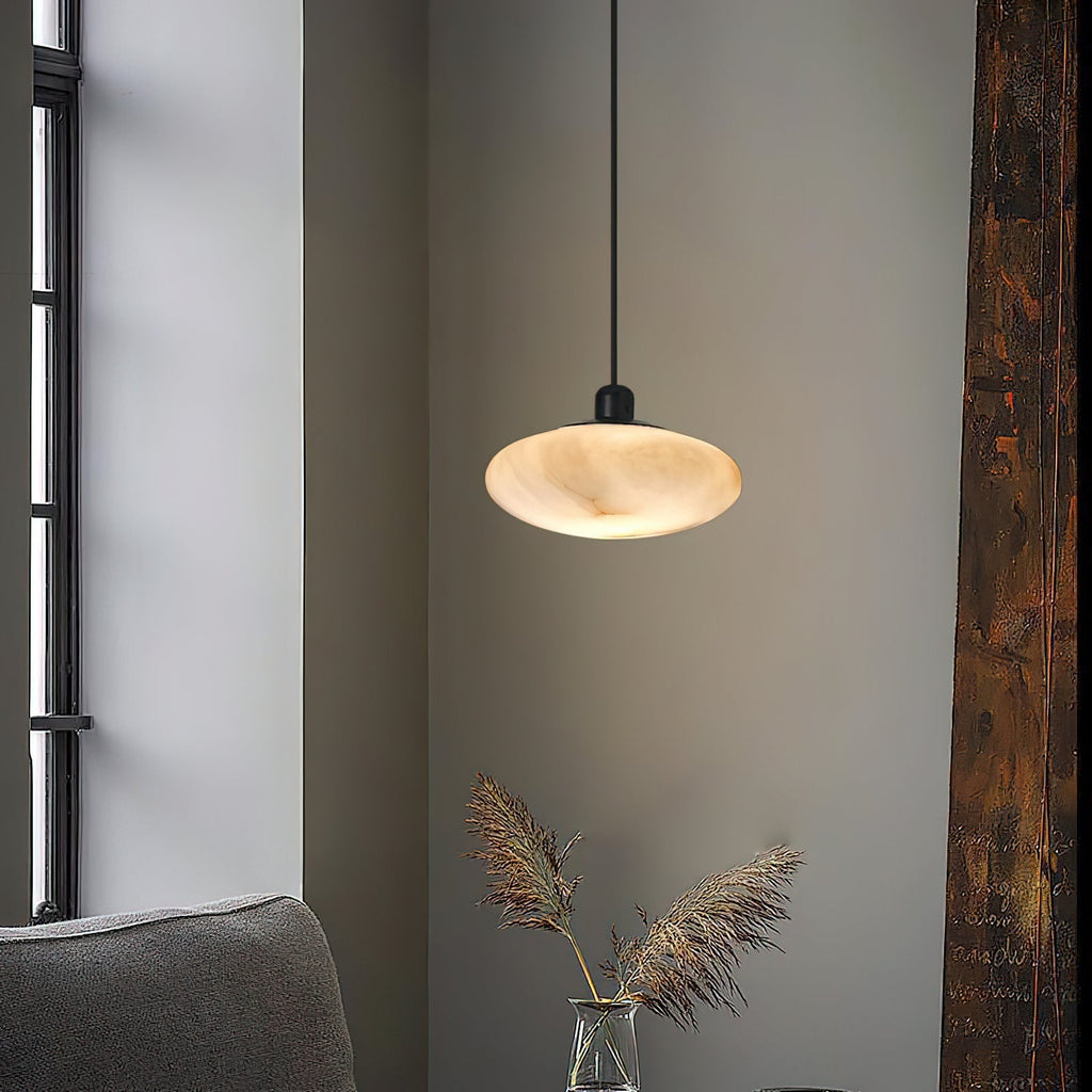 Clérachic Mini Discus pendant light in a corner by a tall window, glowing above dried pampas grass in a glass vase.