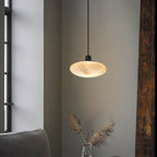 Clérachic Mini Discus pendant light in a corner by a tall window, glowing above dried pampas grass in a glass vase.