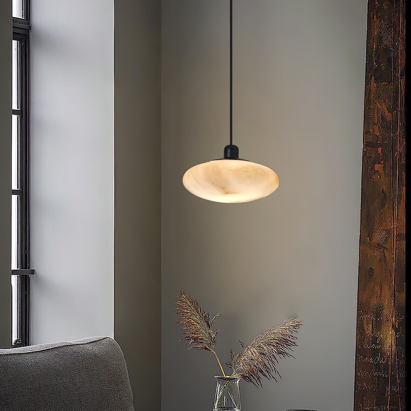 Clérachic Mini Discus pendant light in a corner by a tall window, glowing above dried pampas grass in a glass vase.