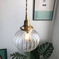 Clérachic Mini Glass Pendant Light in fluted clear glass, lit, hanging beside framed prints and a monstera leaf.