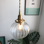 Clérachic Mini Glass Pendant Light in fluted clear glass, lit, hanging beside framed prints and a monstera leaf.