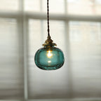 Clérachic Mini Glass Pendant Light in Blackish Green, lifted to a clear aqua by the daylight through the blind behind it.