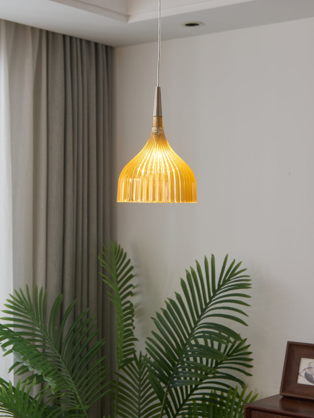 Clérachic fluted pendant light in amber, lit against the curtain wall with the palm fronds spreading well below the shade.