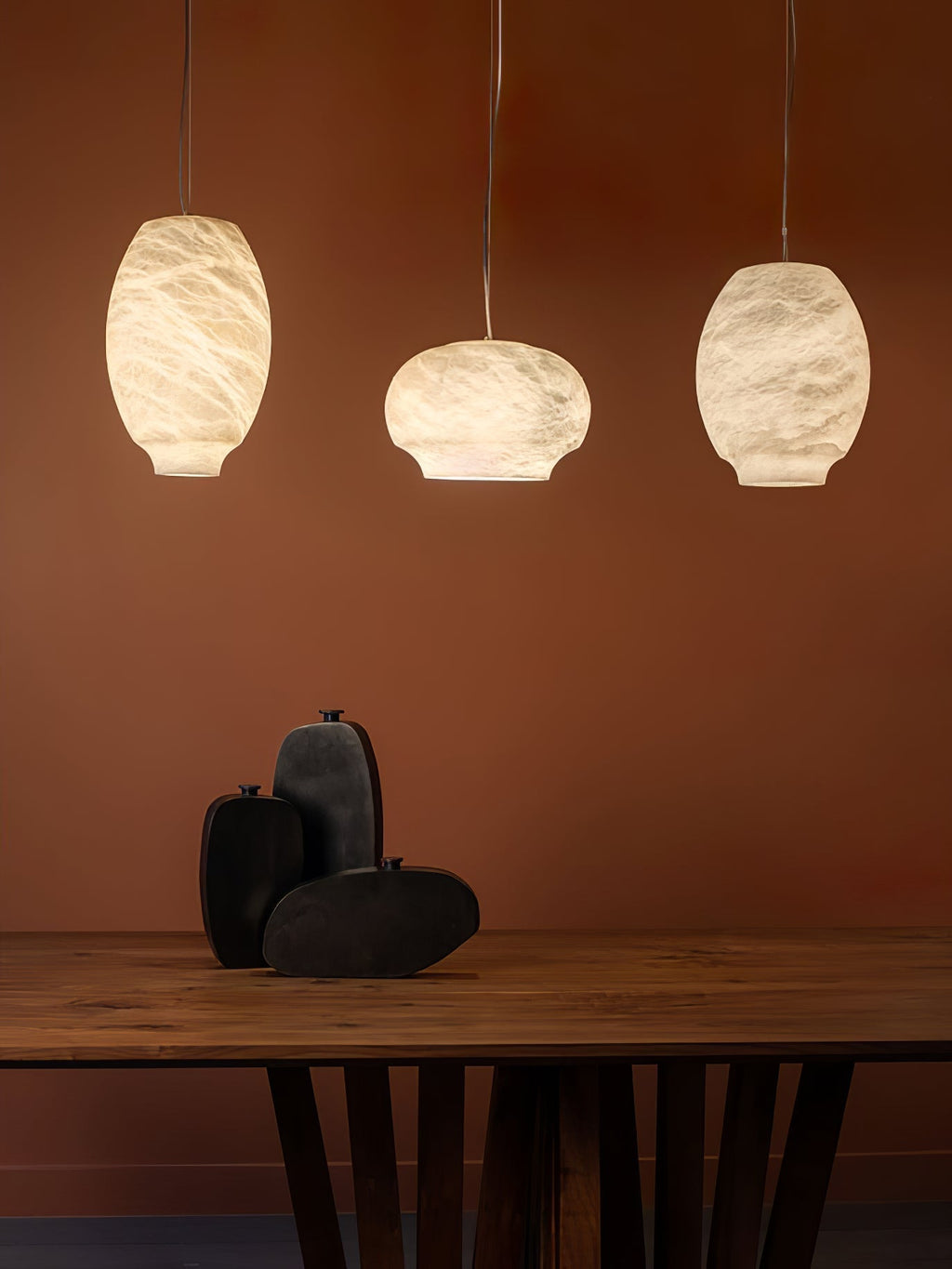 Three Clérachic alabaster pendants in mixed shapes above a timber table on a rust-red wall.
