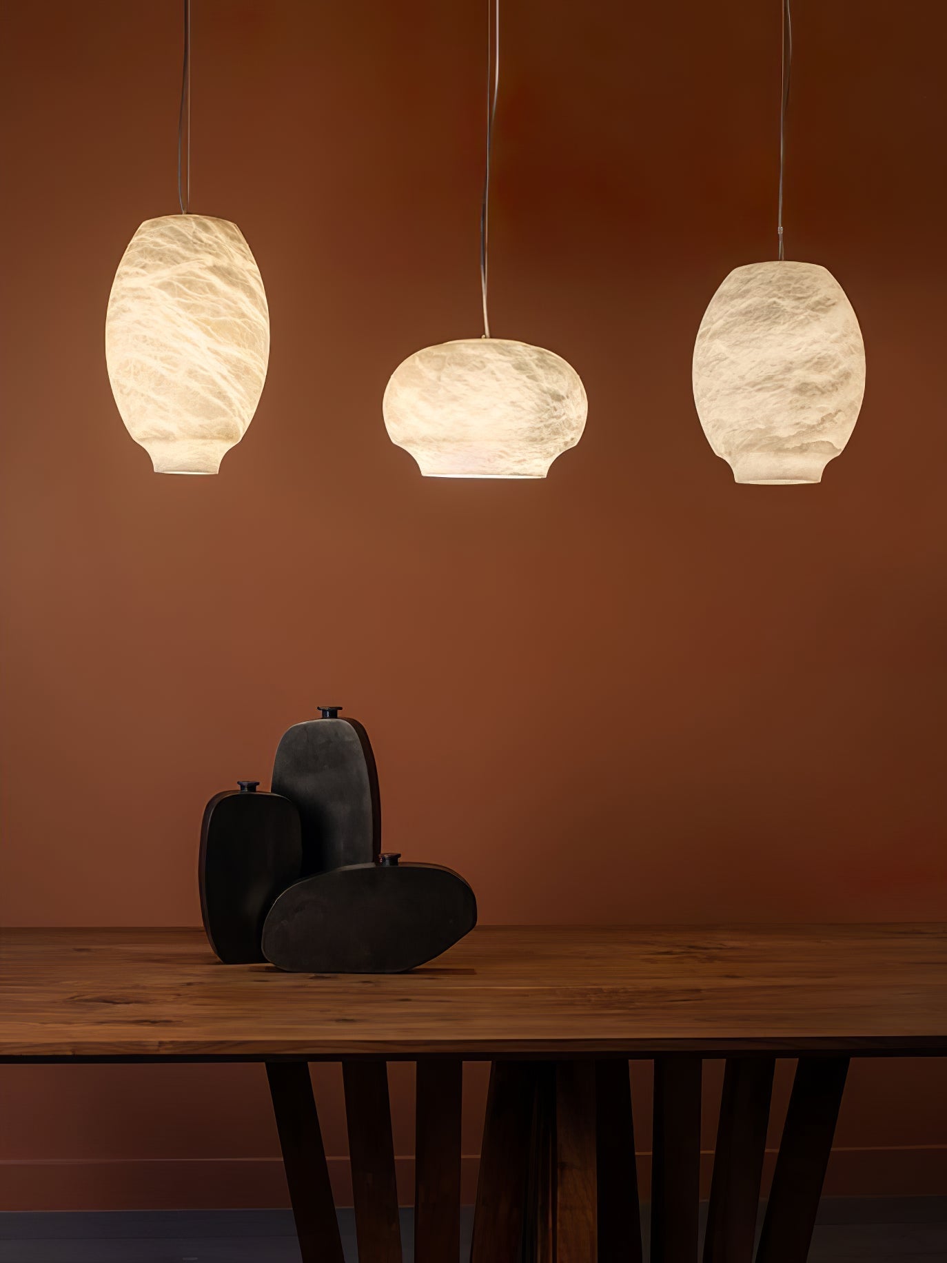 Three Clérachic alabaster pendants in mixed shapes above a timber table on a rust-red wall.