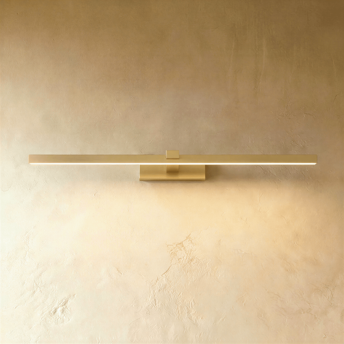 Clérachic slim brushed gold linear wall light in its longest length, lit along the diffuser on a textured limewash wall.
