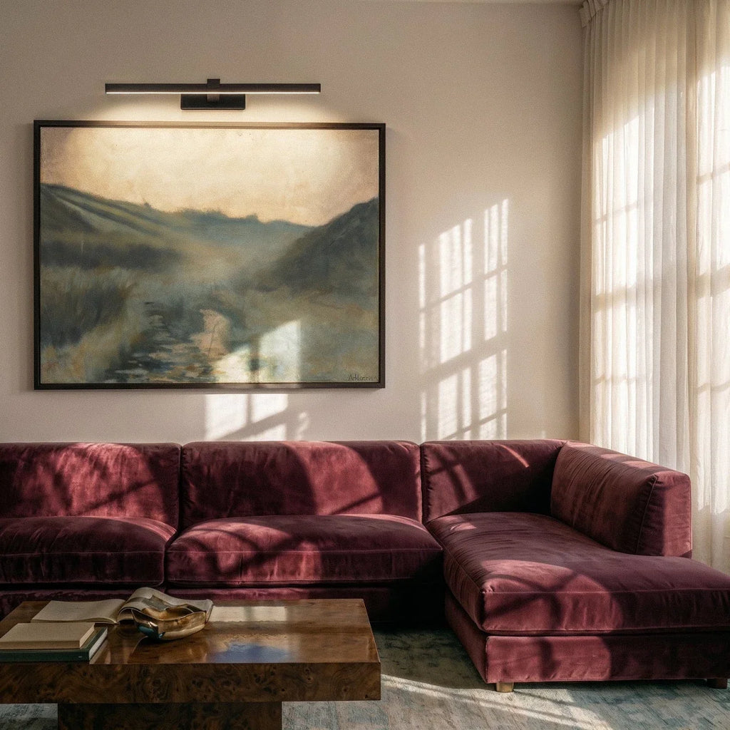 Clérachic black linear picture-style wall light above a large landscape painting, over a plum velvet sofa in a sunlit room.