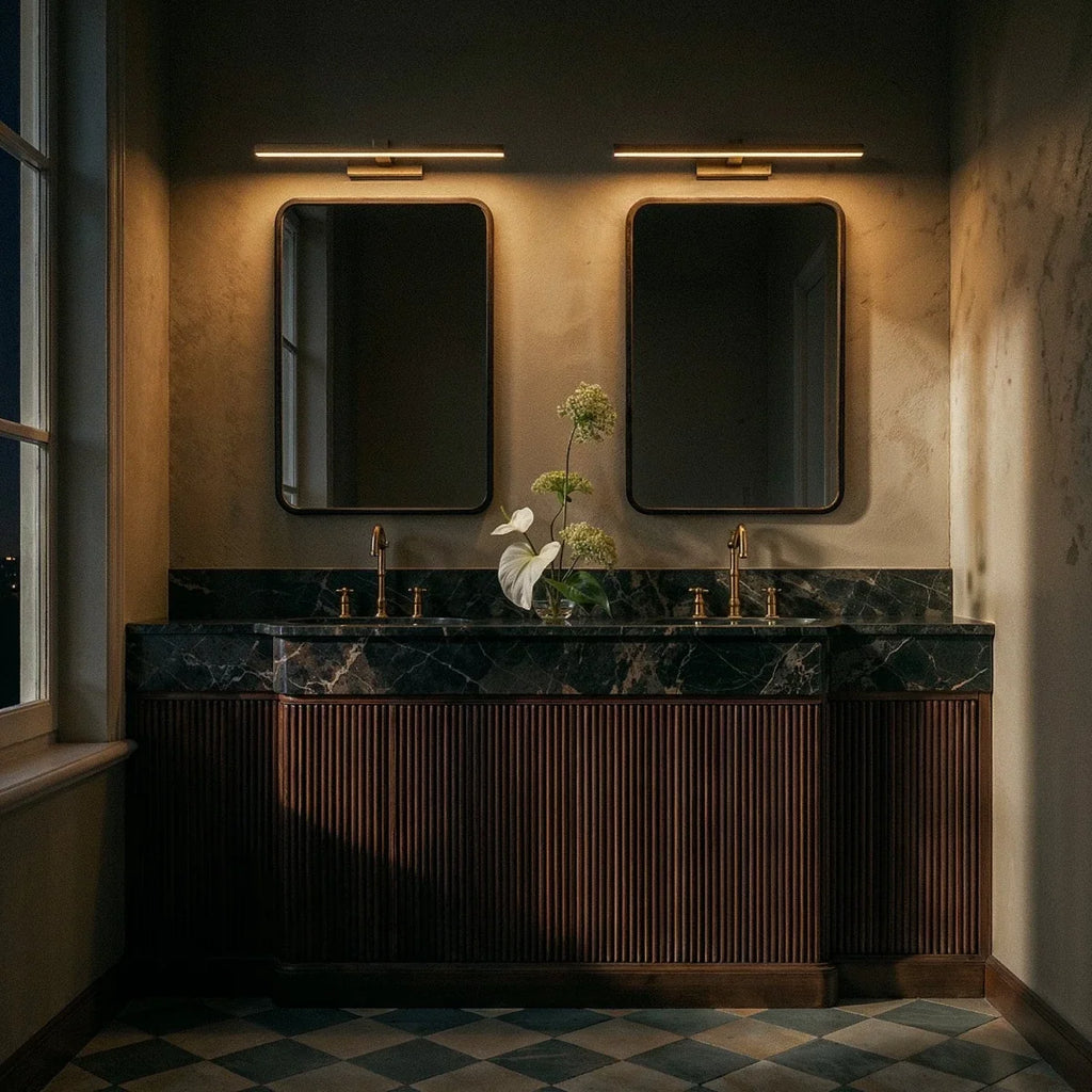 Clérachic gold linear wall lights glowing above two mirrors at night, over a marble vanity with fluted wood cabinets.