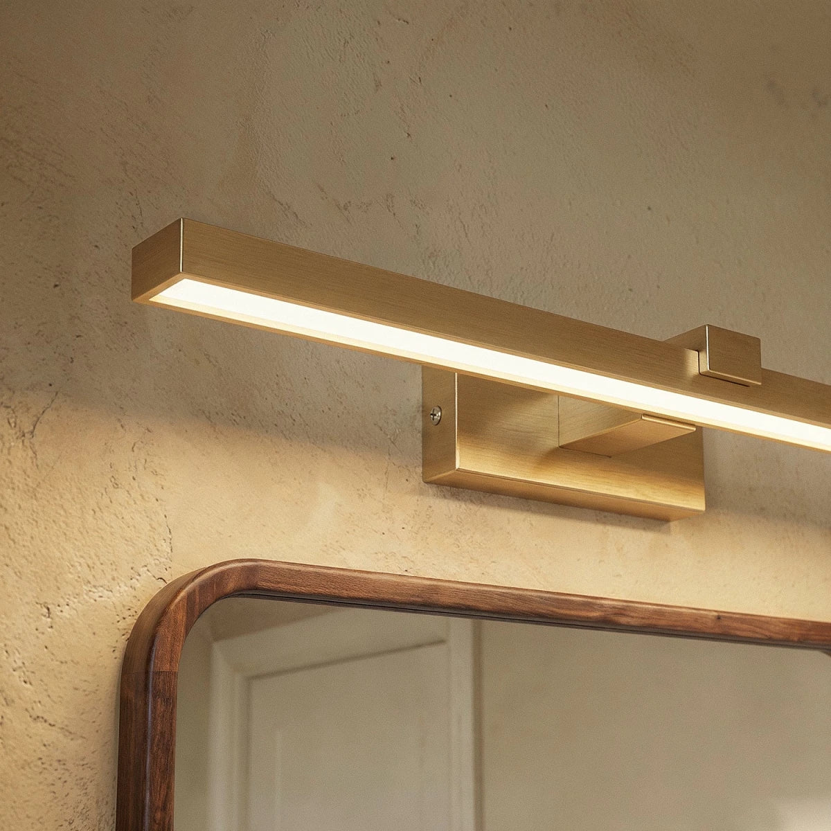 Close view of a Clérachic brushed gold linear wall light, its diffuser glowing above a wood-framed mirror on a plaster wall.