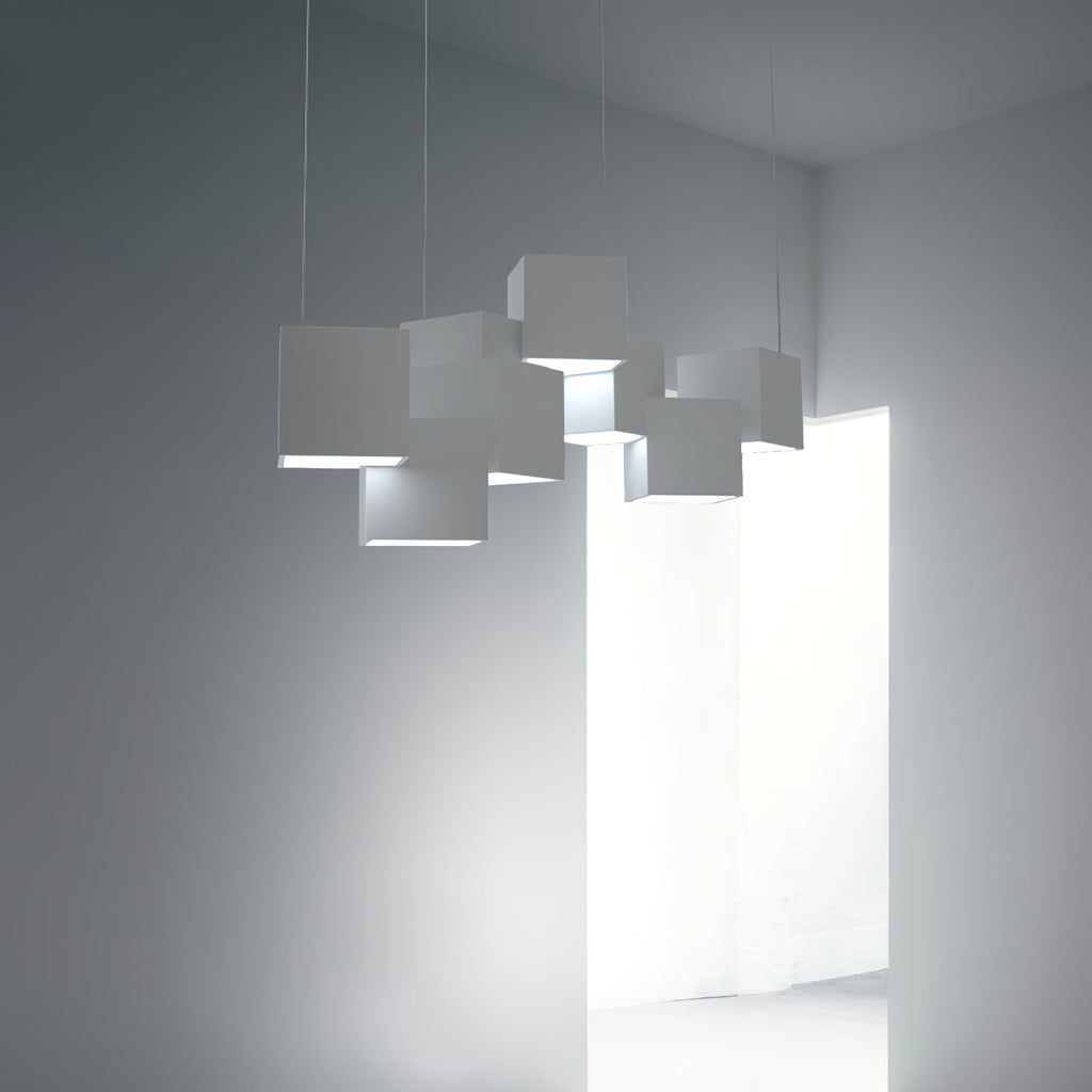 Clérachic cube chandelier in white, lit below the cubes, in a grey room with an open doorway to a bright space behind.