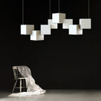 A Clérachic chandelier of white cubes over a dark wall, with a wooden chair draped in white cloth below it.
