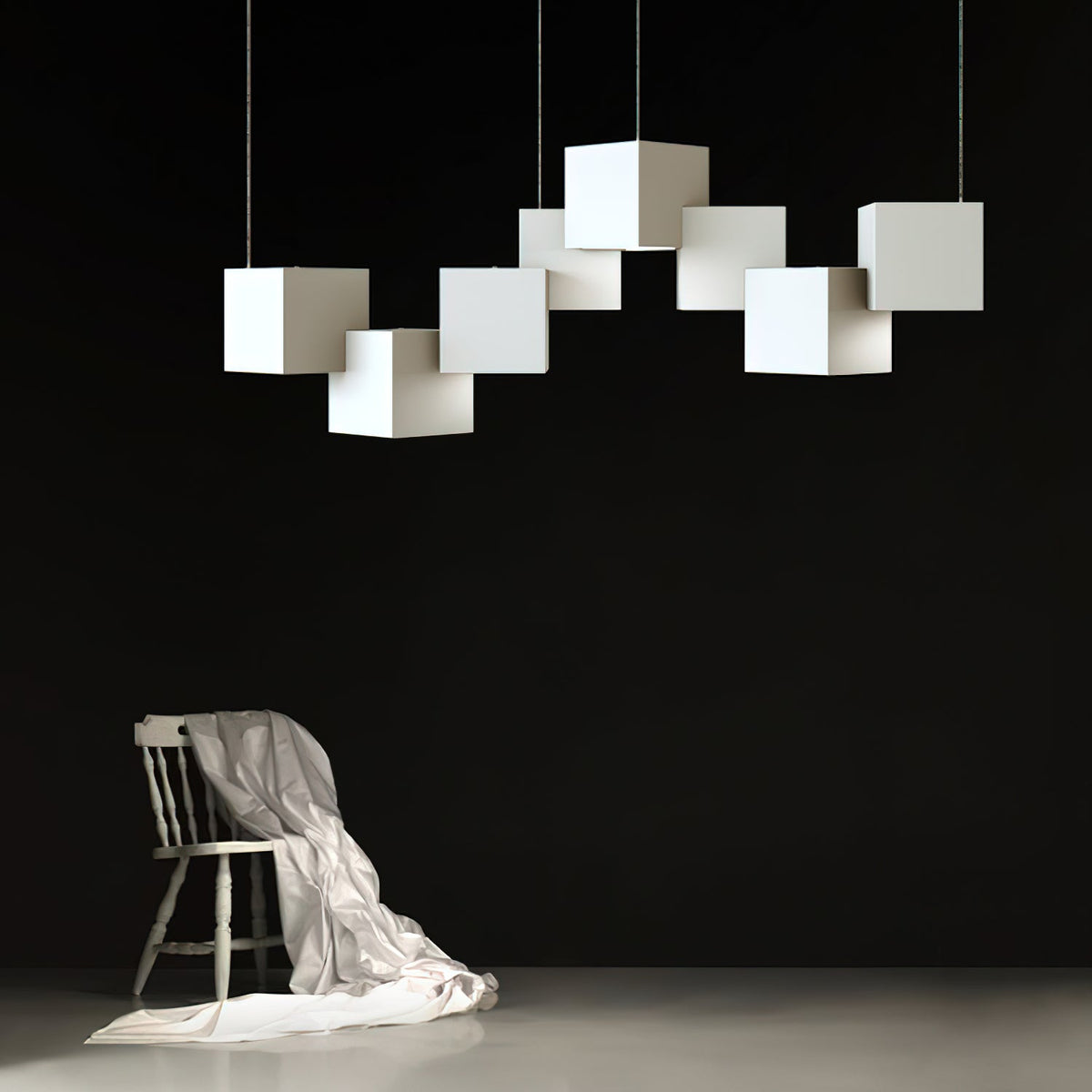 A Clérachic chandelier of white cubes over a dark wall, with a wooden chair draped in white cloth below it.