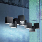 Black Clérachic cube chandelier in front of a blue-green glass and concrete wall, with light glowing from beneath each cube.