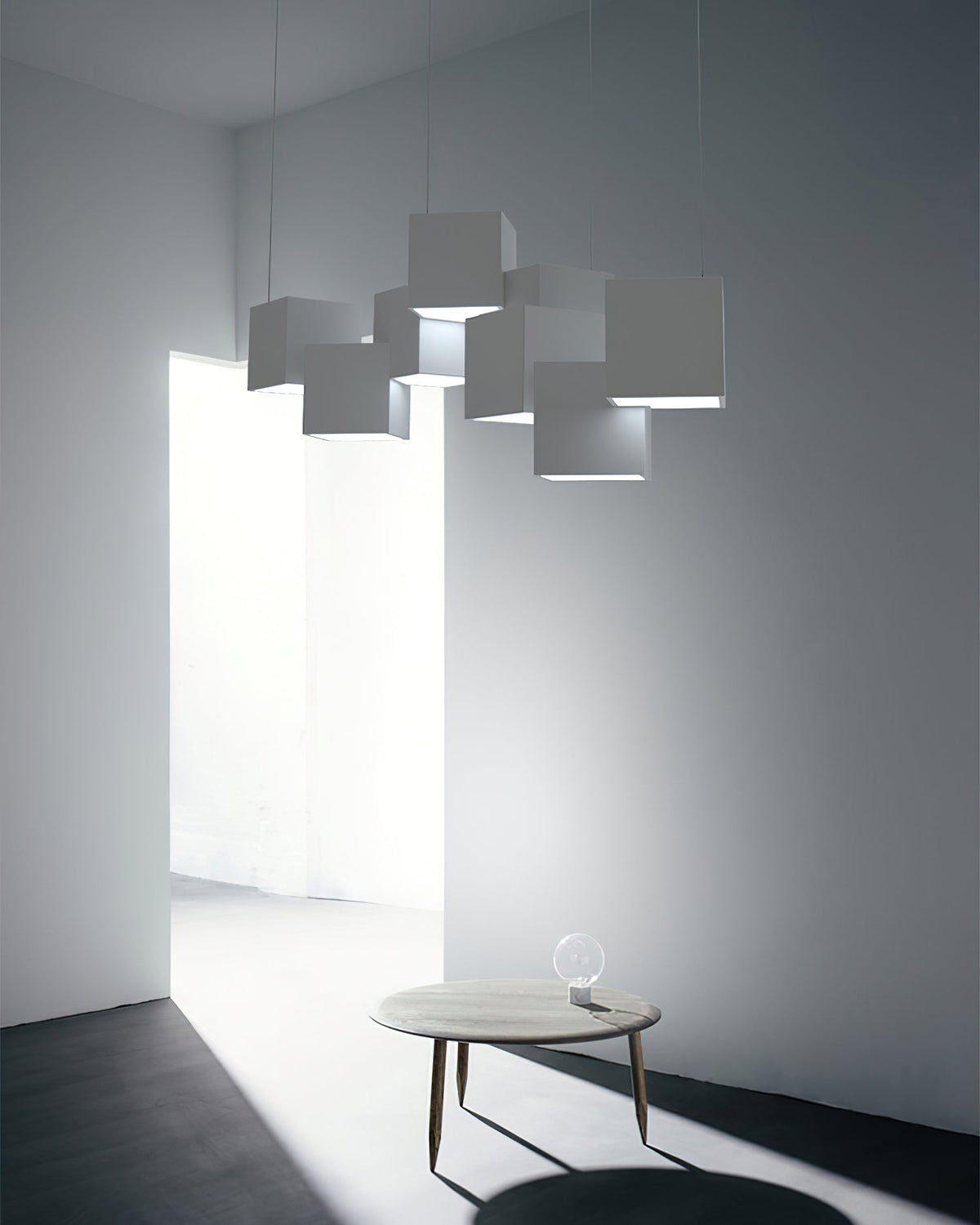 A Clérachic white cube chandelier in a grey room with a doorway and a round wooden coffee table with a bulb on it.