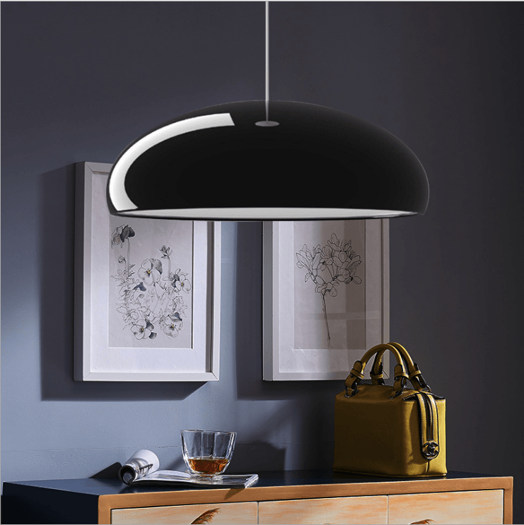 Glossy black Clérachic dome pendant lit from below, hanging over a wooden sideboard with framed botanical prints and a mustard handbag.