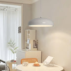 A light grey Clérachic dome pendant above a small round table in a bright corner, with a white shelf unit and a potted fern behind.