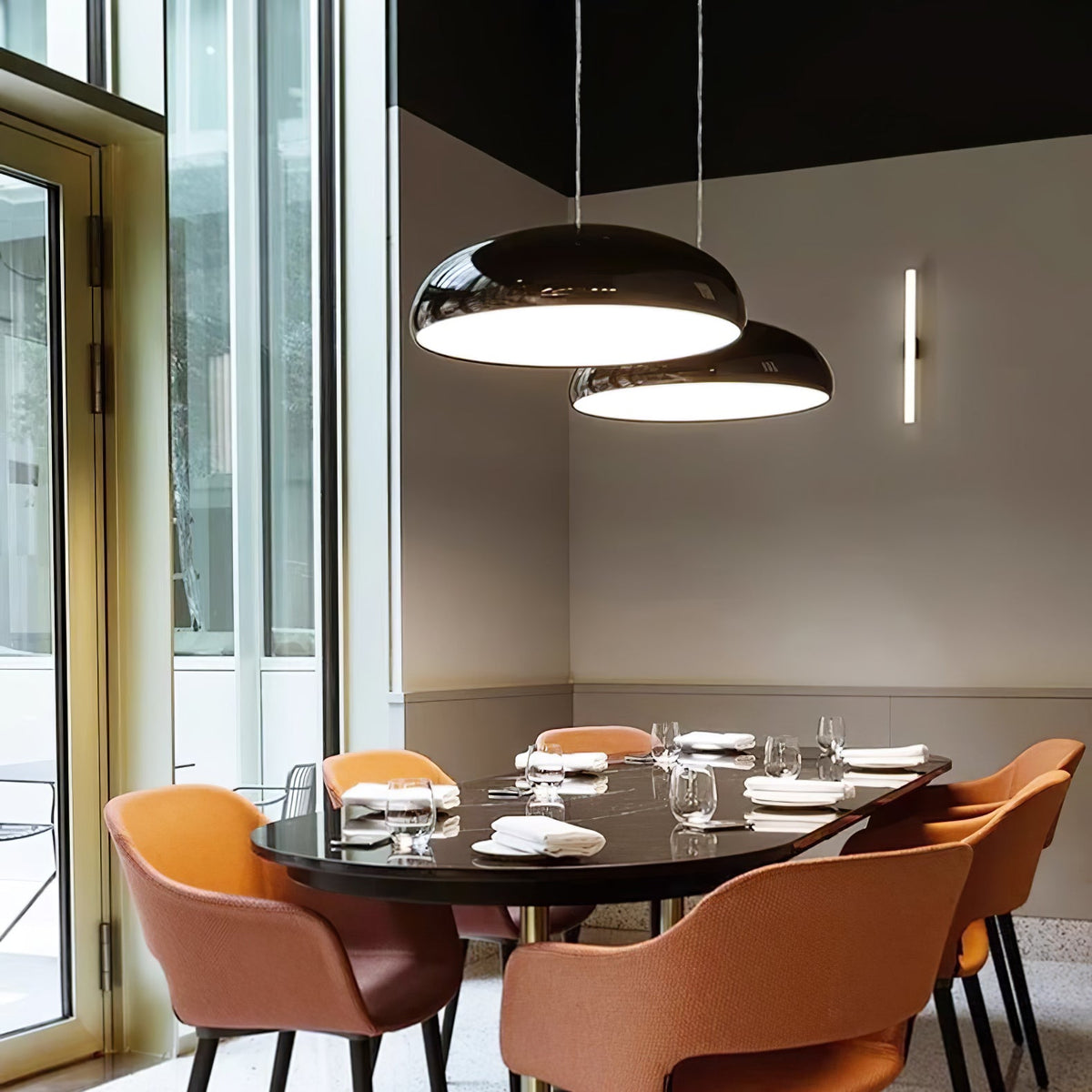 Two black Clérachic dome pendants at different heights over a dark oval dining table, with tan armchairs and tall windows beyond.
