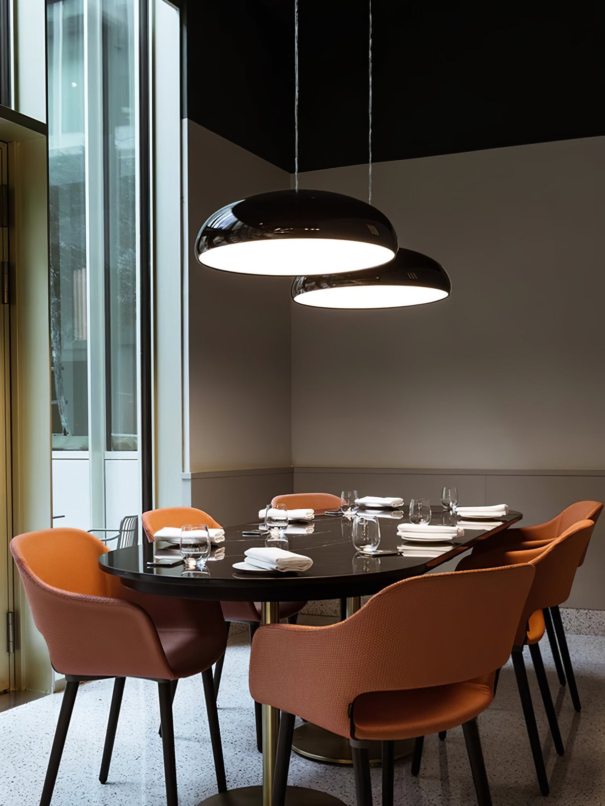 Pair of black Clérachic dome pendants glowing above a dining table in a restaurant-style room, with orange chairs pulled in around it.
