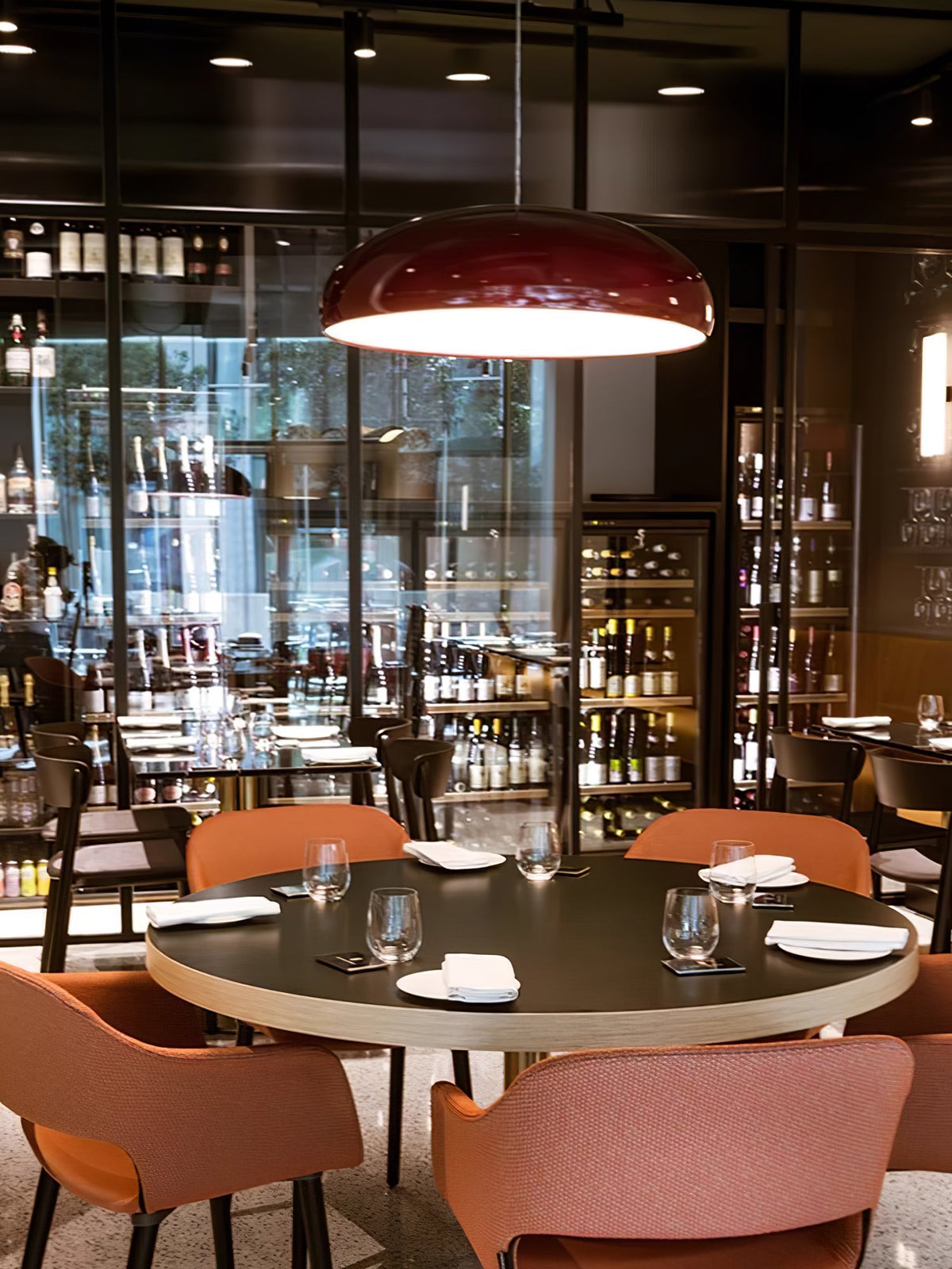 Dark-red Clérachic pendant lit above a round table in a busy wine bar, with a glass-fronted bottle wall behind it.