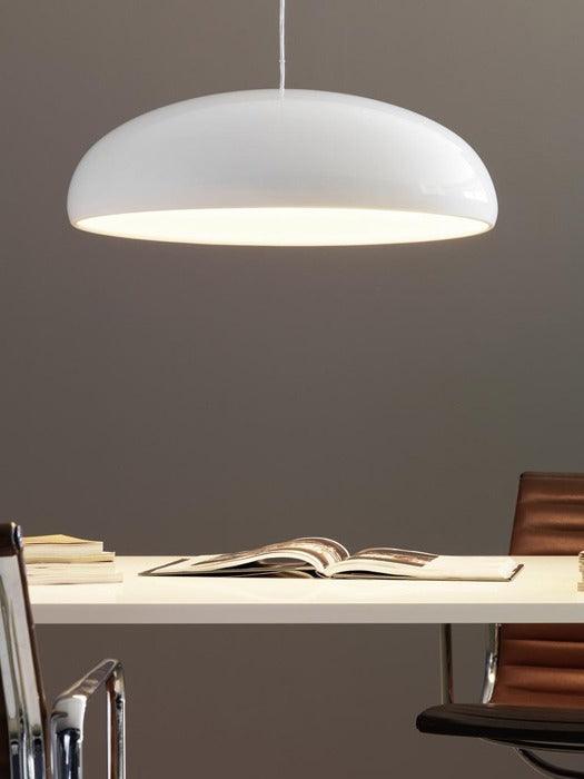A white Clérachic dome pendant on a taupe wall, with its warm underside glow falling toward a white desk and an open book.