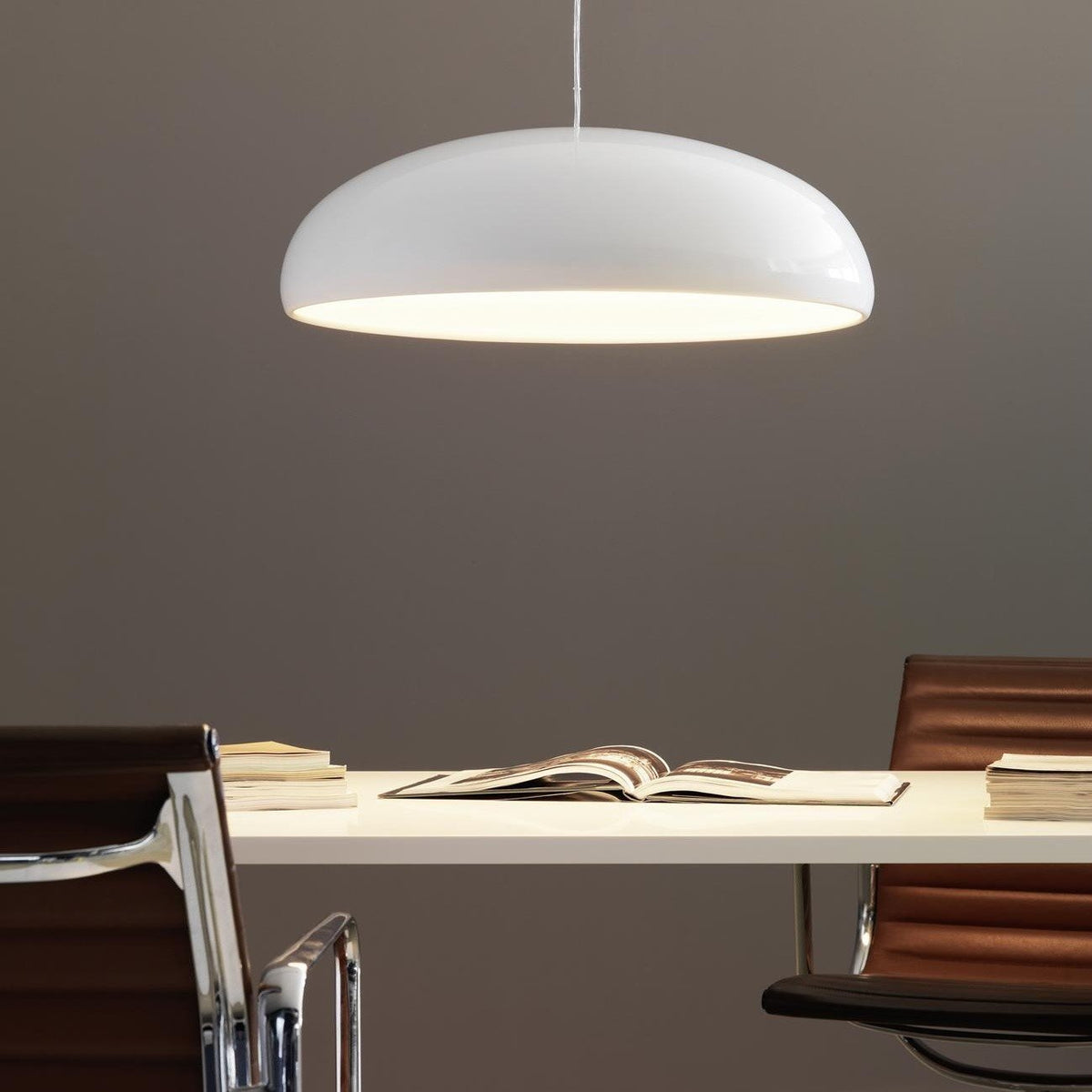 White Clérachic dome pendant glowing warmly over a long white desk, with books and two leather chairs on a taupe wall.