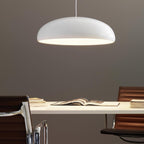 A Clérachic white dome pendant lit warmly at its rim, hanging over a cream desk with an open book and leather chairs.