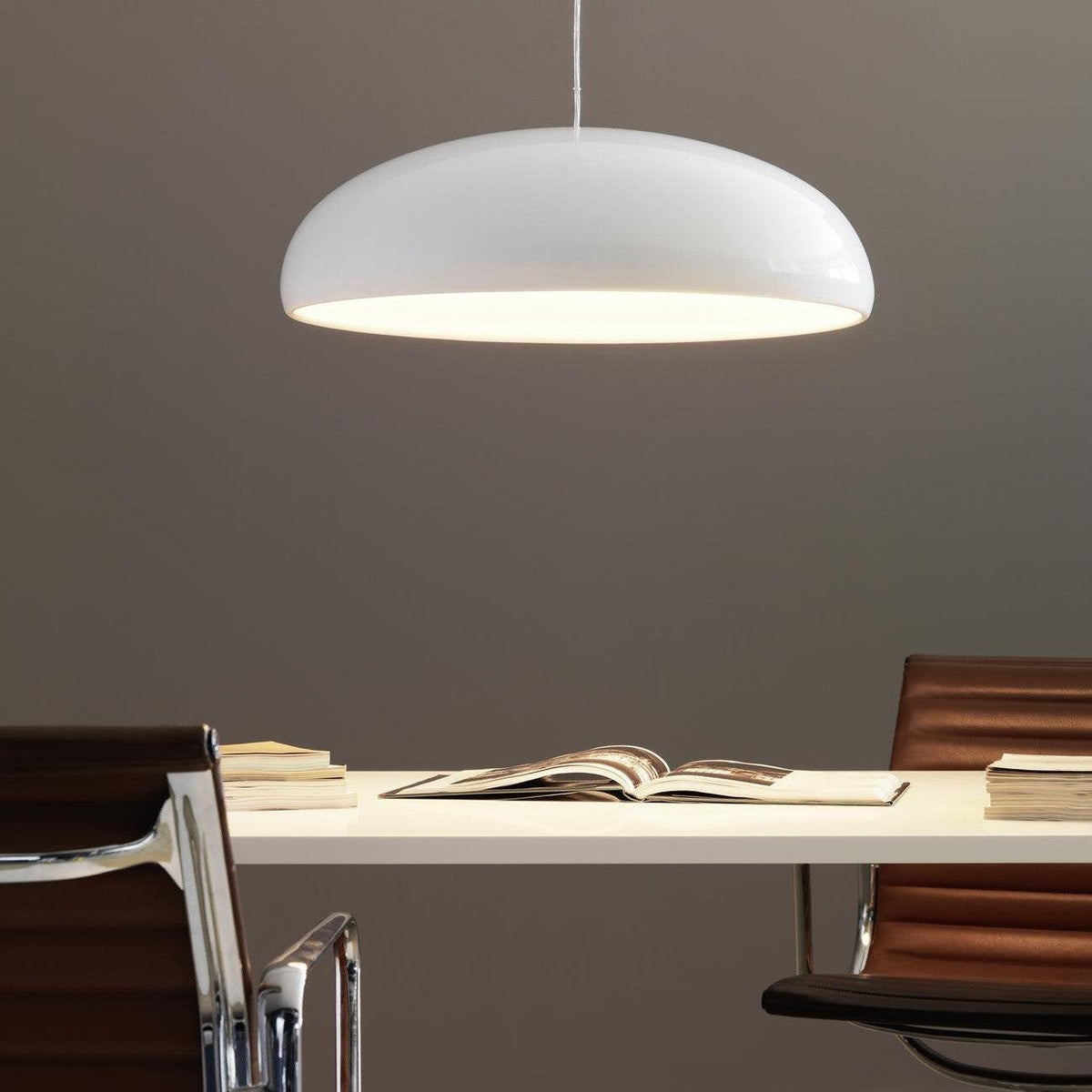 A Clérachic white dome pendant lit warmly at its rim, hanging over a cream desk with an open book and leather chairs.