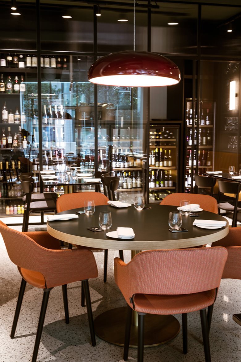 Dark-red Clérachic dome pendant glowing high above a round dining table, with salmon-coloured chairs and a lit bar behind.