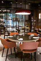 Dark-red Clérachic dome pendant glowing high above a round dining table, with salmon-coloured chairs and a lit bar behind.