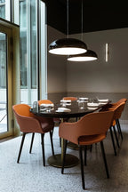 Two black Clérachic pendants lit above a dark dining table, with orange chairs, a speckled floor and a tall window beside.