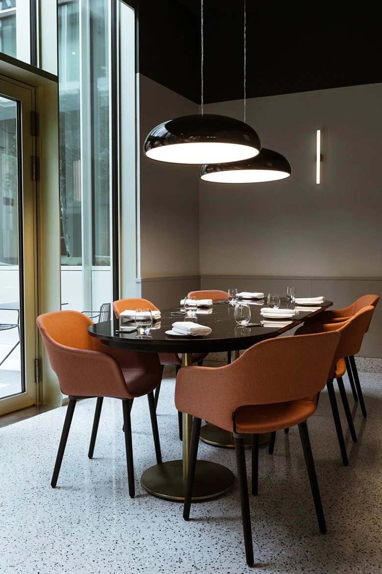 Two black Clérachic pendants lit above a dark dining table, with orange chairs, a speckled floor and a tall window beside.