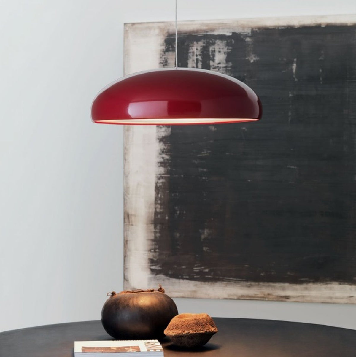 Dark-red Clérachic dome pendant in front of a large charcoal and cream canvas, with two clay pots on a round table below.