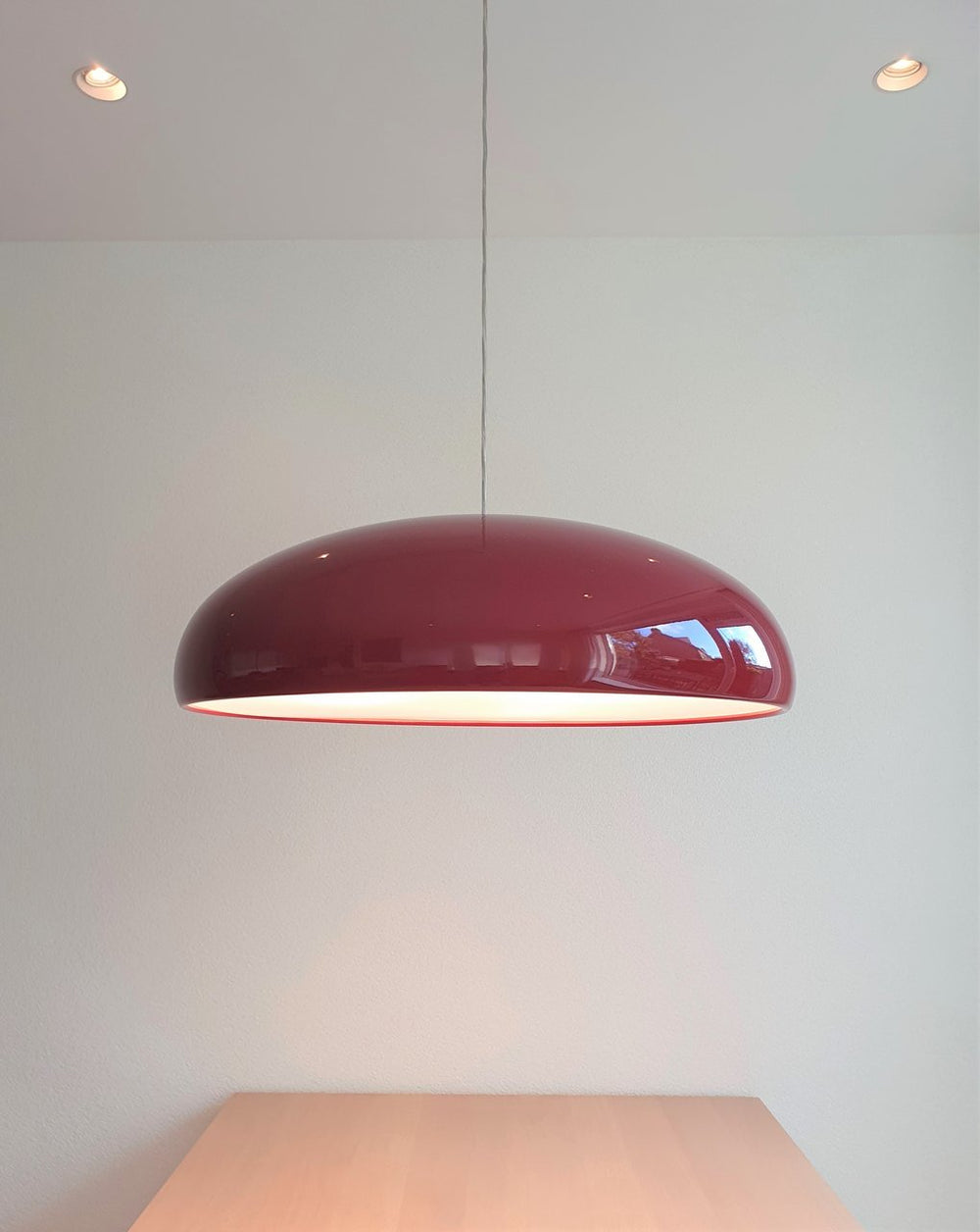 Dark-red Clérachic dome pendant hung high on a pale wall above a light wooden table, with spotlights in the ceiling.