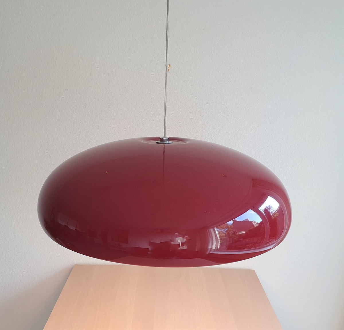 Close view of a glossy dark-red Clérachic dome pendant, its curved shell reflecting the room, above a pale wooden table.