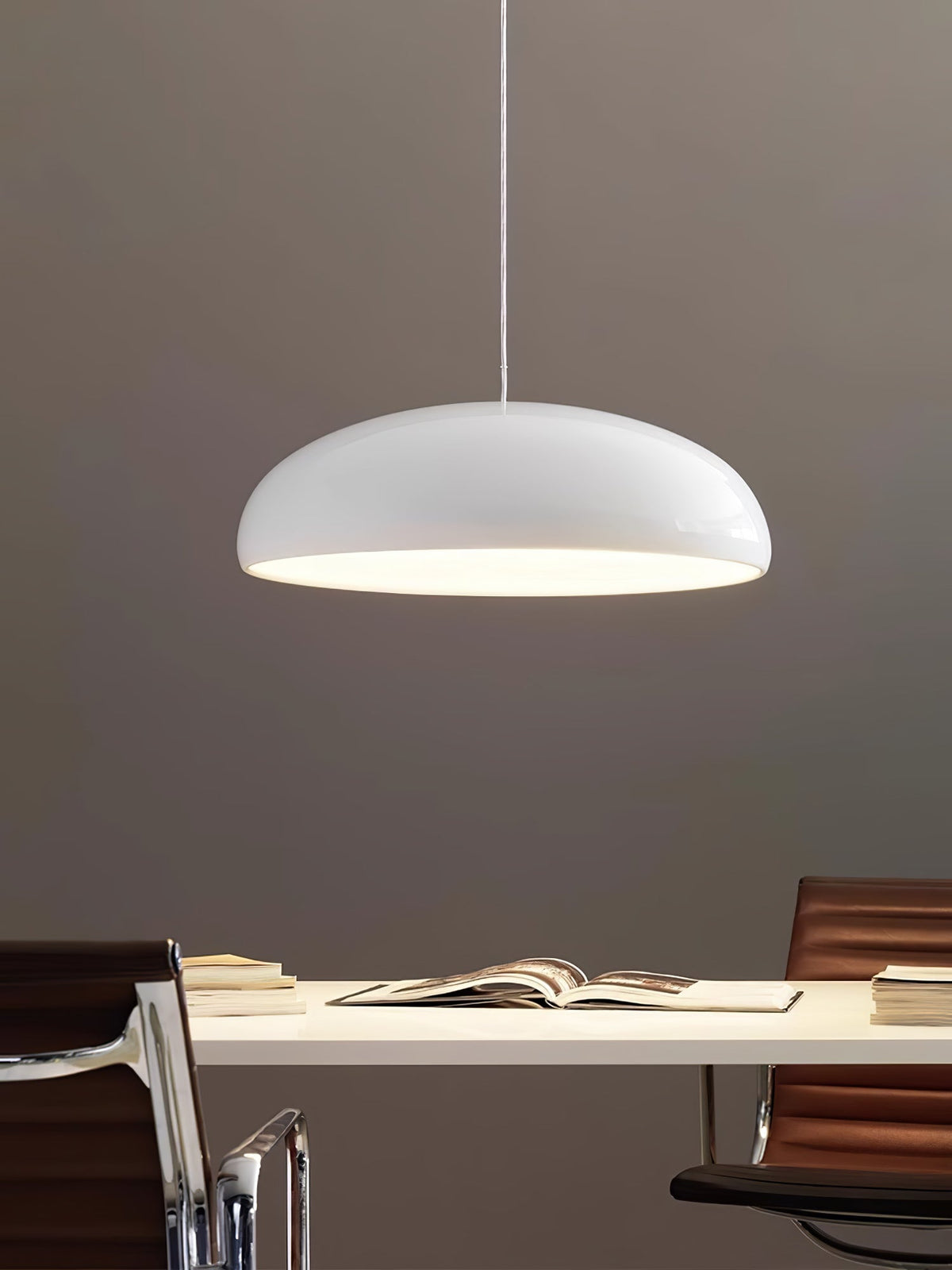 White Clérachic dome pendant lit warmly from below, above a white desk with an open book against a taupe wall.