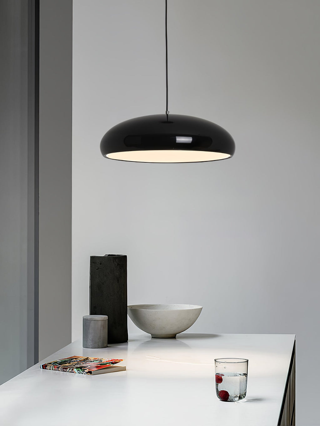 Black Clérachic dome pendant against a pale wall, above a white table with a stone bowl, a dark vase and a glass of water.