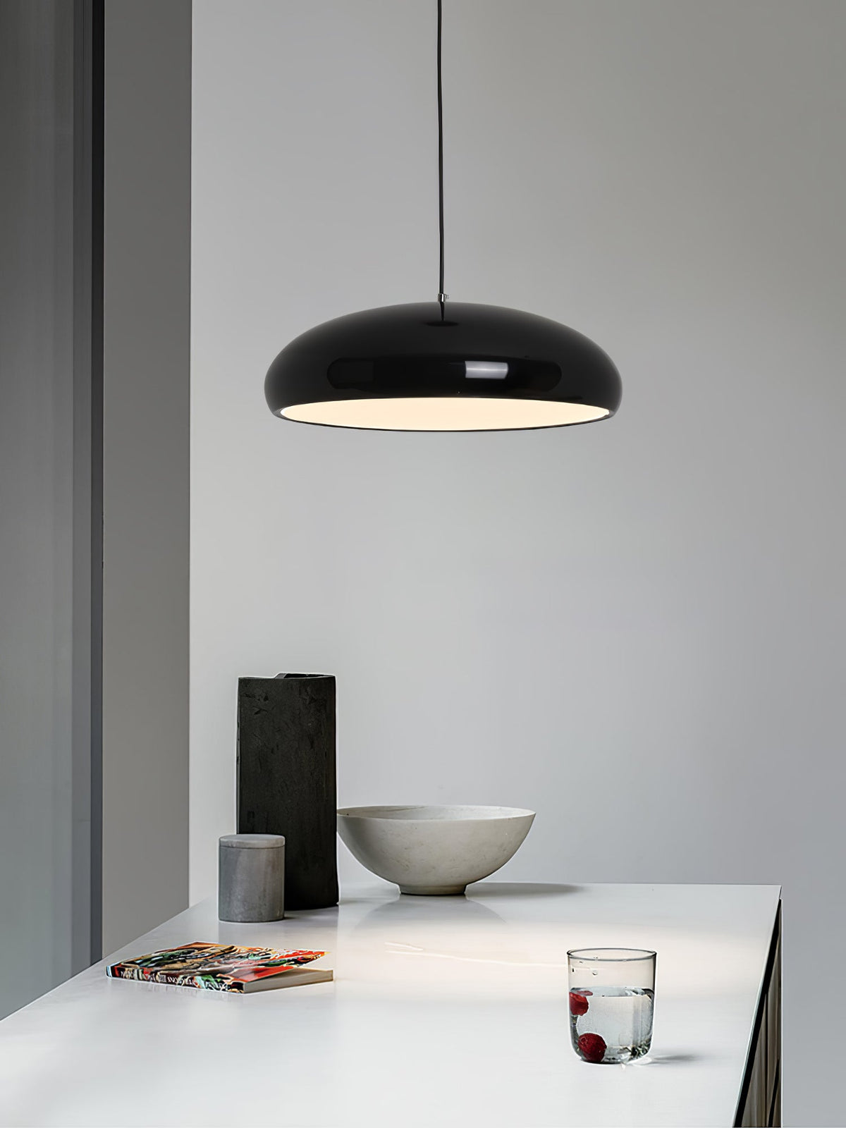 Black Clérachic dome pendant against a pale wall, above a white table with a stone bowl, a dark vase and a glass of water.
