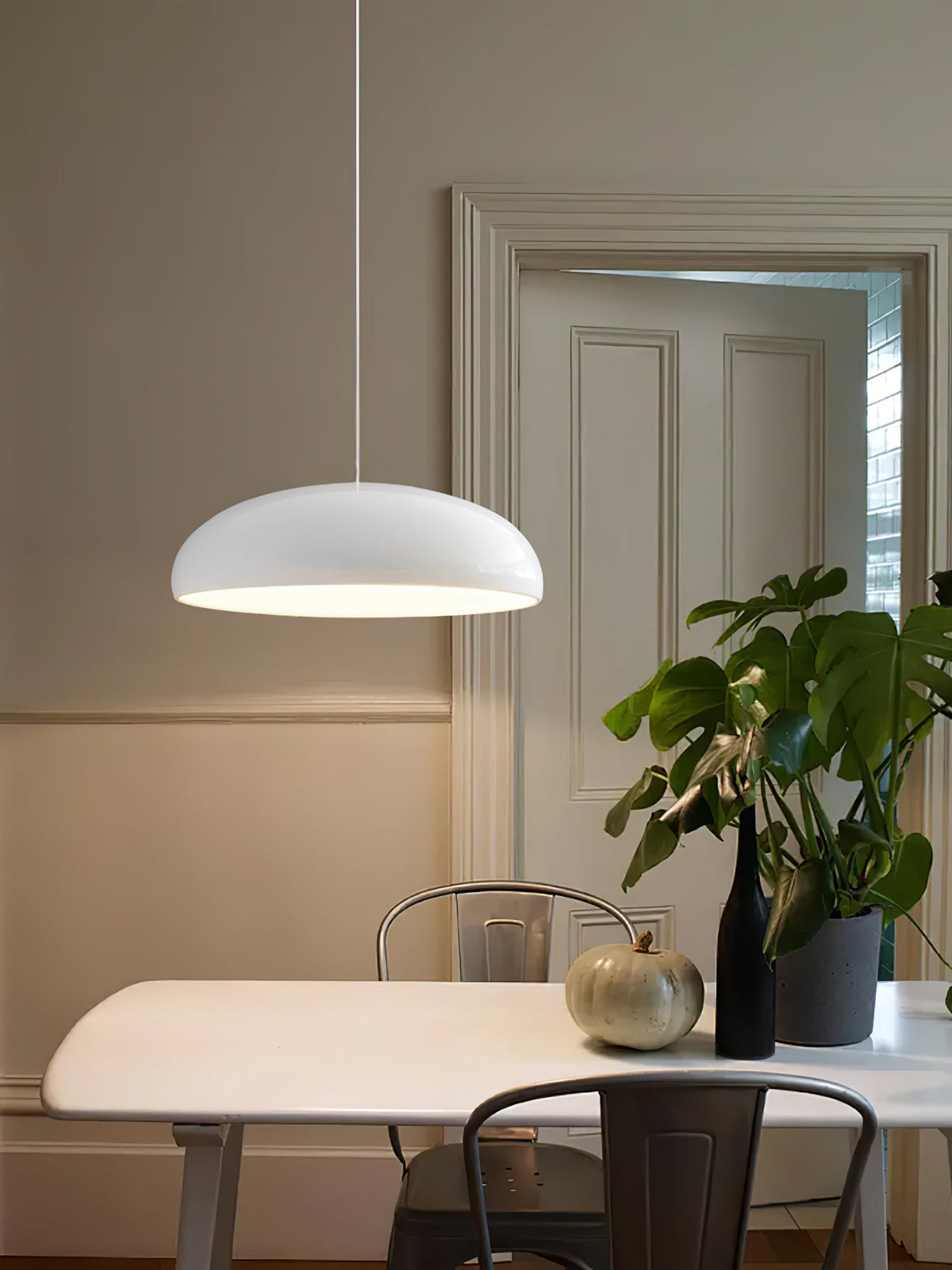 A white Clérachic dome pendant in a room with panelled doors, hung above a white table with a monstera plant and a pumpkin.