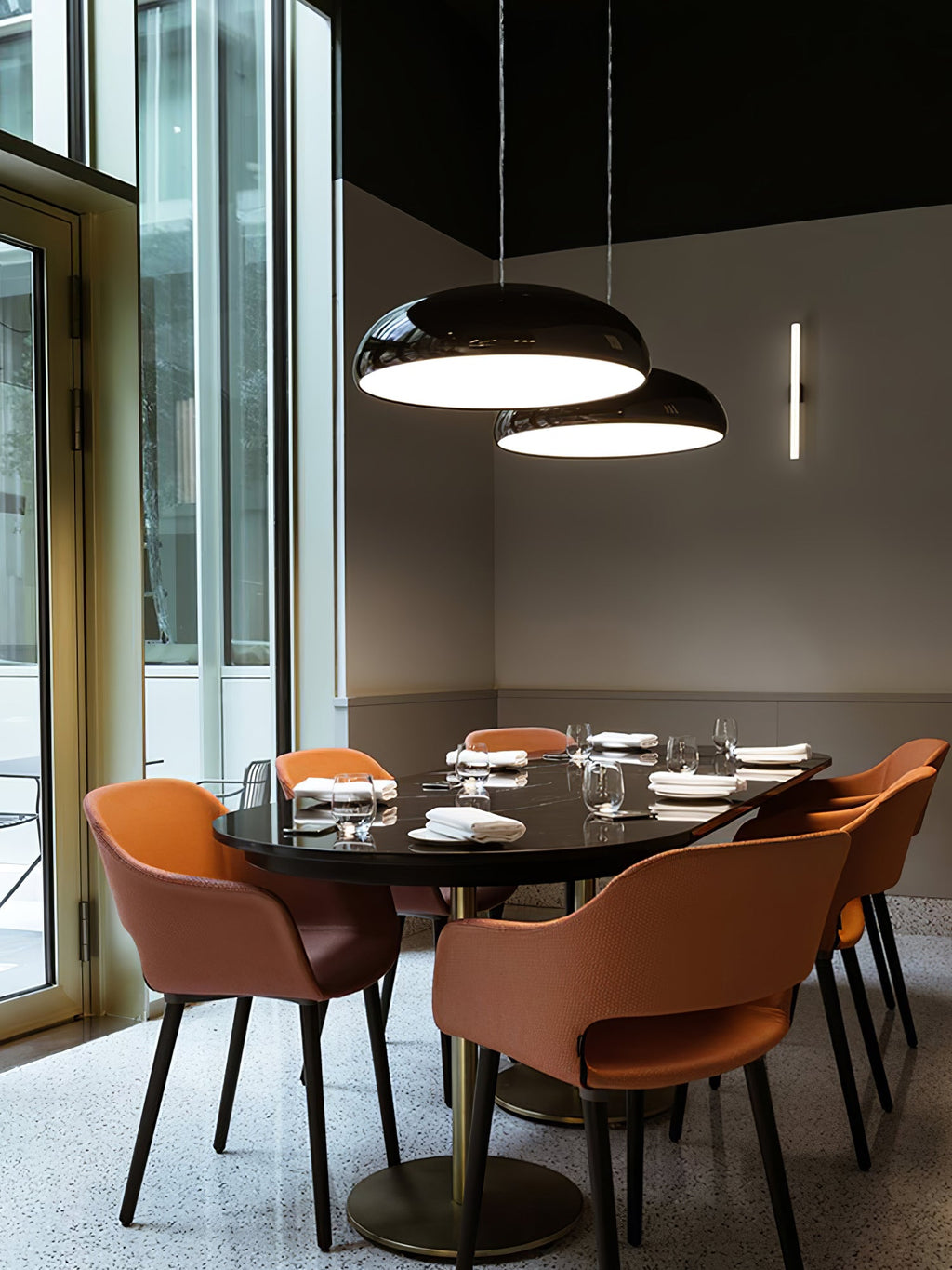Two black Clérachic dome pendants above a dark oval table, with orange chairs and floor-to-ceiling windows behind.