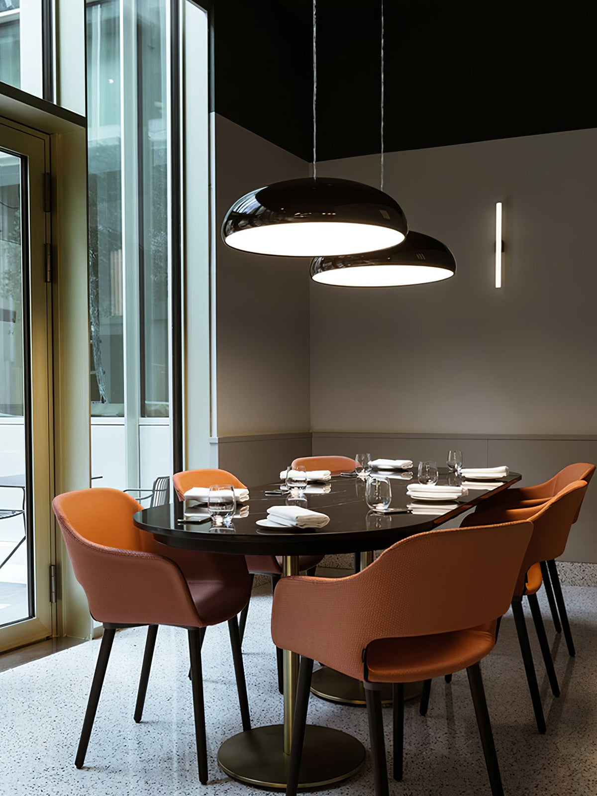 Two black Clérachic dome pendants above a dark oval table, with orange chairs and floor-to-ceiling windows behind.