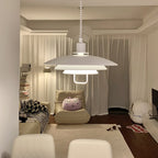 A lit white Clérachic pendant hangs in a living room, its layered shades glowing warm above a cream sofa and white chairs.