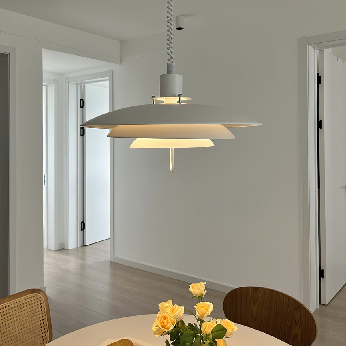 Above a round dining table with yellow roses, a white Clérachic pendant glows softly, with a bright hallway behind it.